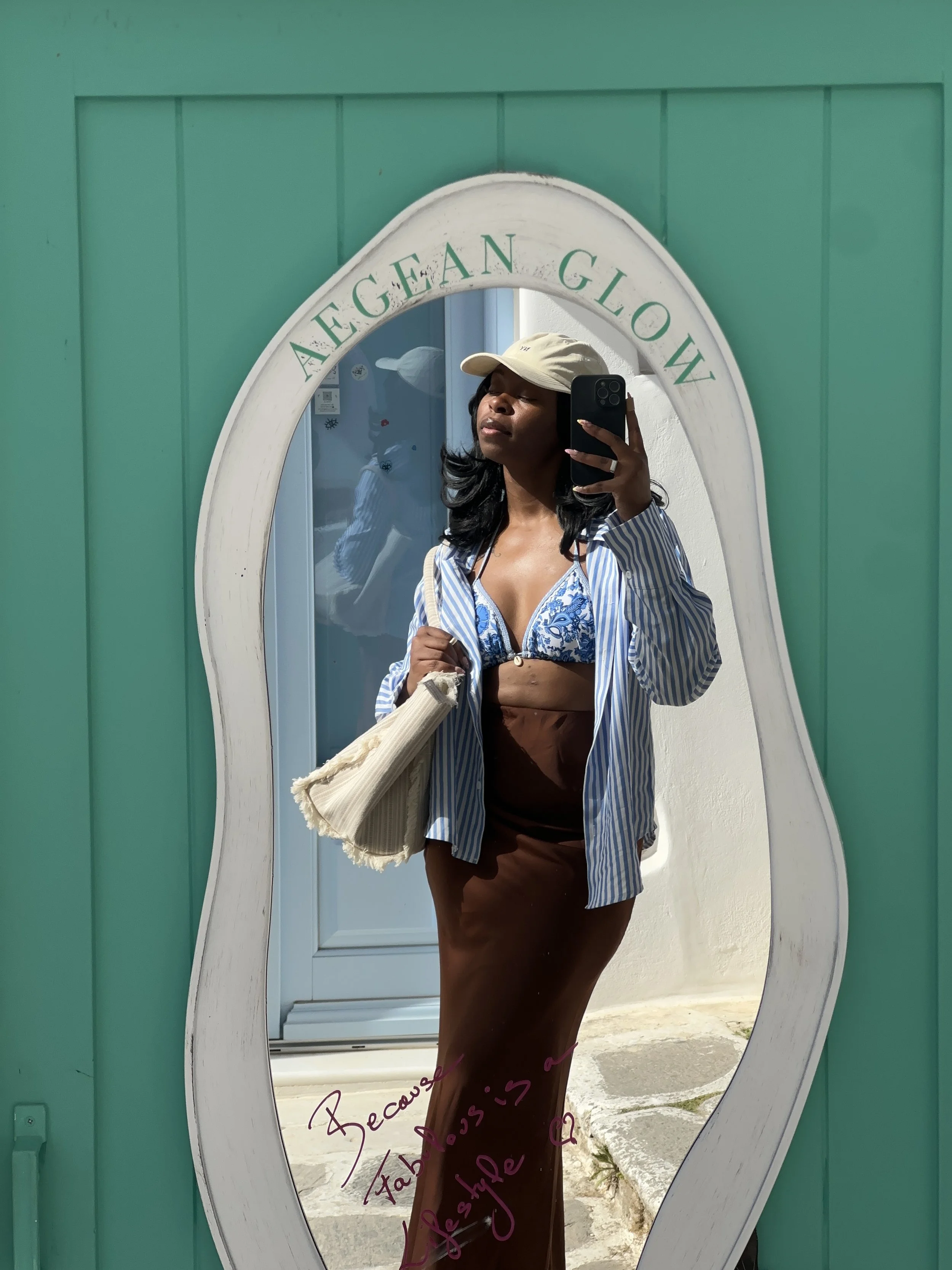 A woman takes a mirror selfie wearing an open blue and white striped button-down shirt over a blue pattern bikini top and a brown high-waisted maxi skirt.