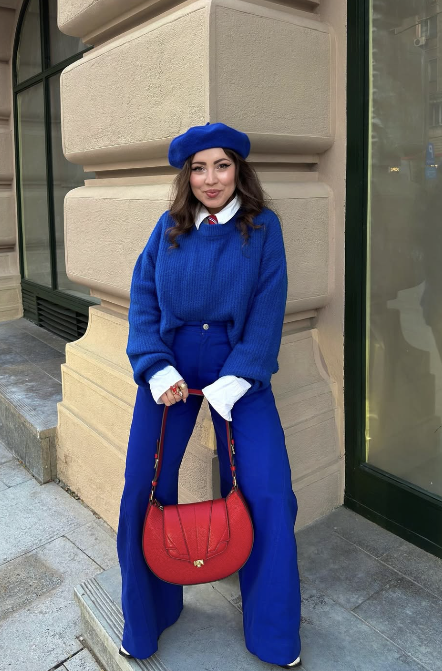 A woman wearing a monochromatic royal blue outfit featuring a fuzzy knit sweater, matching trousers, and a blue beret, accessorized with a bold red handbag.