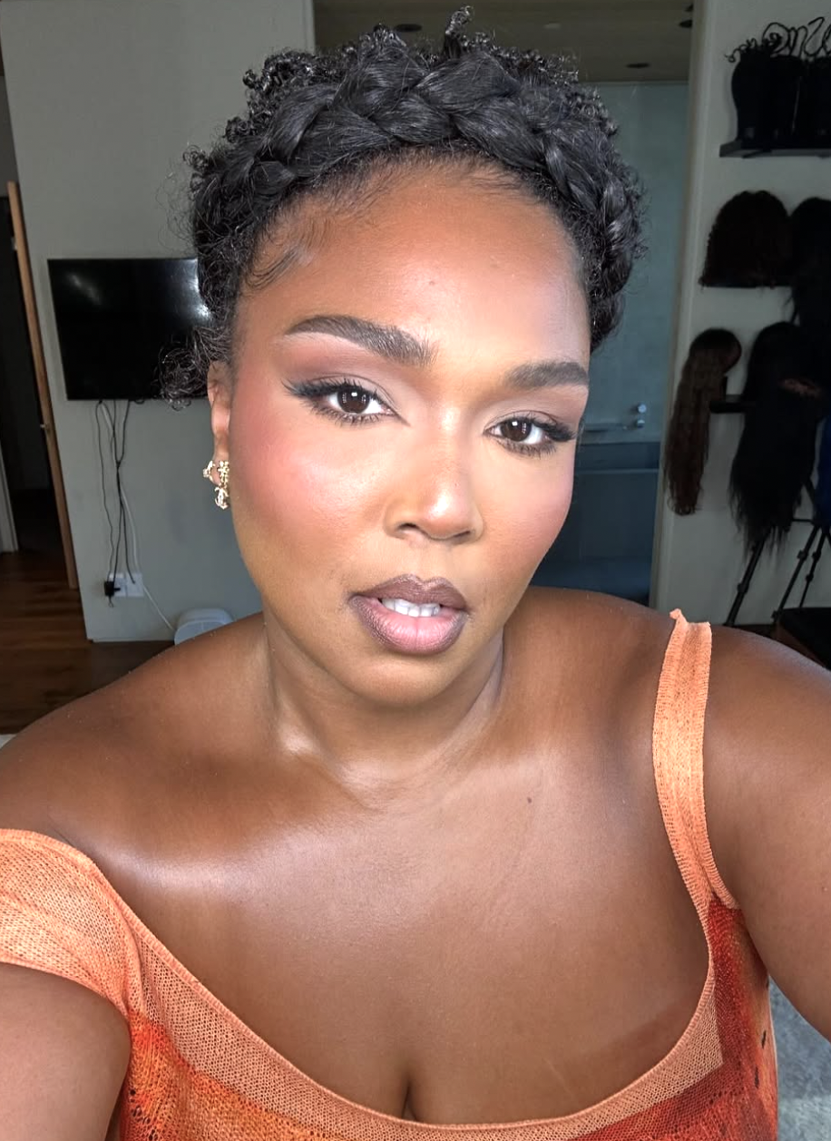 Lizzo wearing an orange off-the-shoulder top, showcasing a high braided crown updo that adds height at the top of the head to elongate a round face.