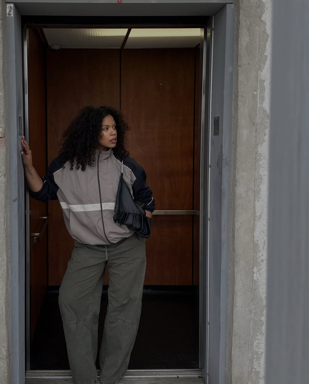 A woman wearing an off-duty utility outfit consisting of an olive green windbreaker, baggy grey cargo pants, and a structured black leather shoulder bag.