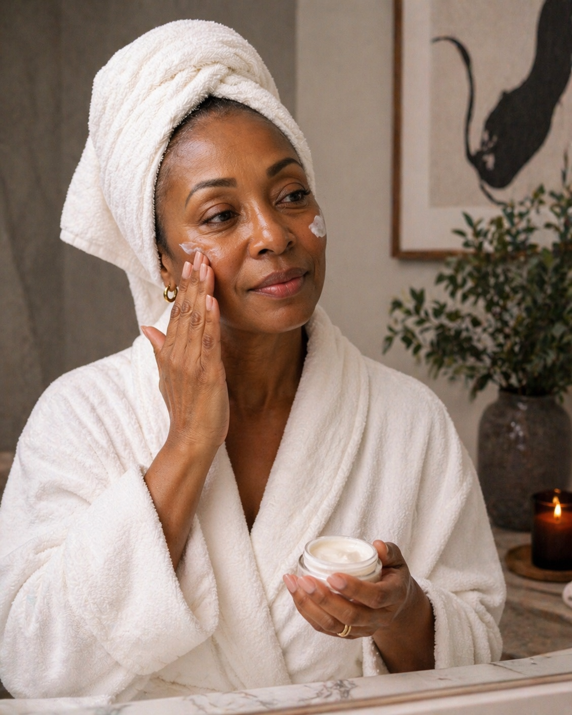 A smiling middle-aged Black woman in a white bathrobe applying face cream to her cheek, representing healthy skincare for women over 50.