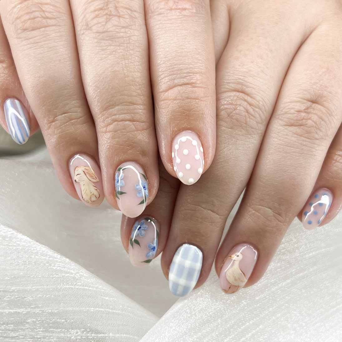 A whimsical mismatched manicure featuring light blue gingham patterns, white polka dots on a cream base, and hand-painted illustrations of a bunny and a chick on almond-shaped nails.