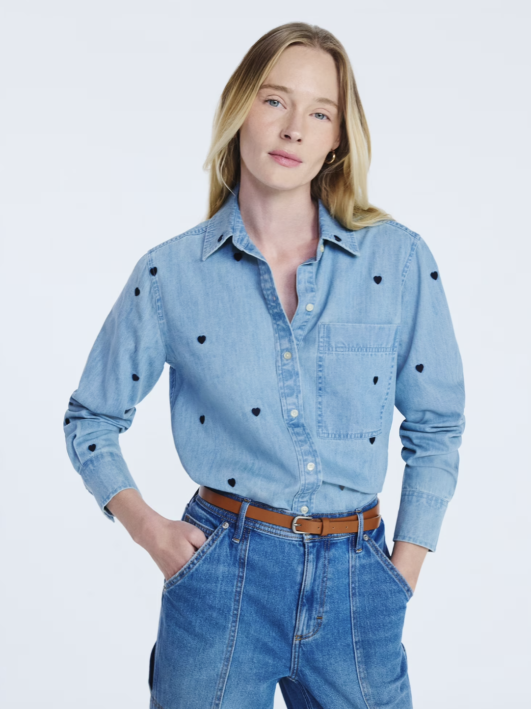 A woman wearing a light-wash denim button-down shirt featuring small black embroidered hearts, tucked into high-waisted blue jeans with a brown leather belt.