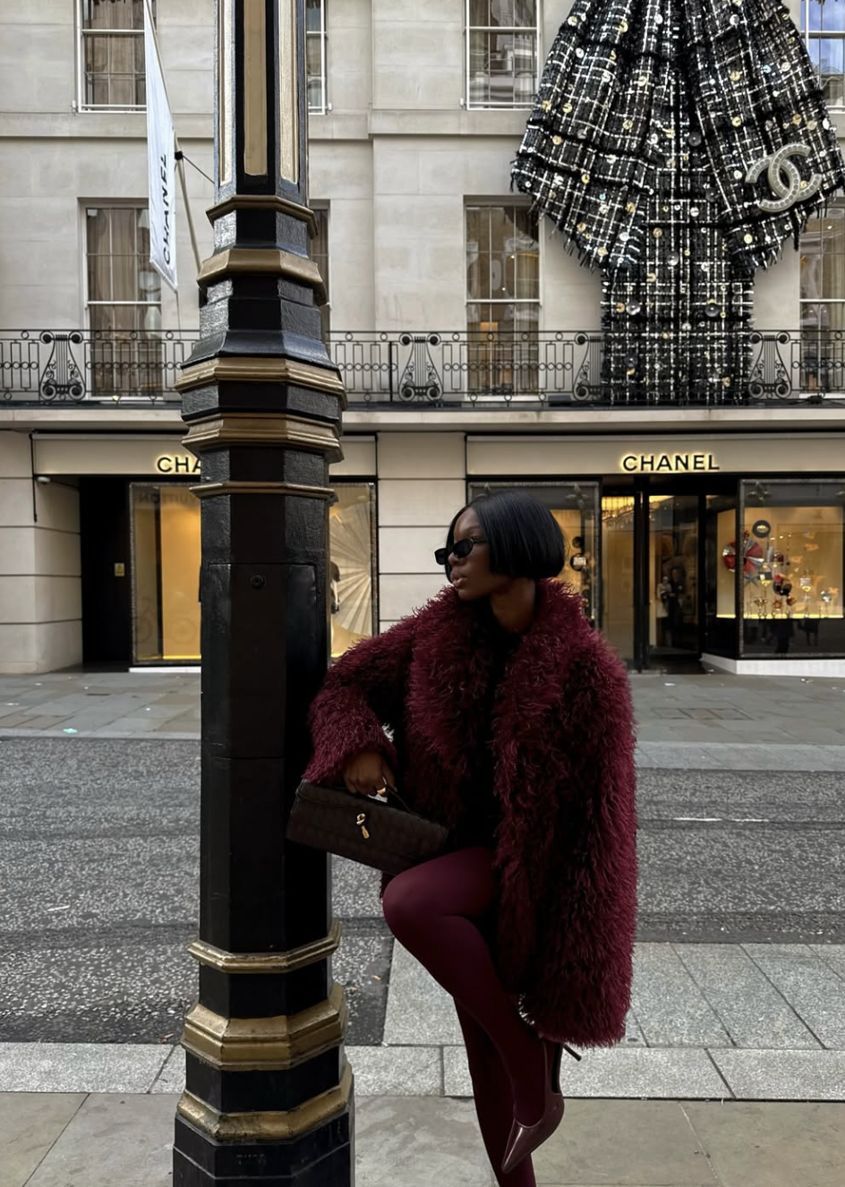 A woman wearing a plush burgundy faux fur coat and black sunglasses standing in front of a Chanel boutique.