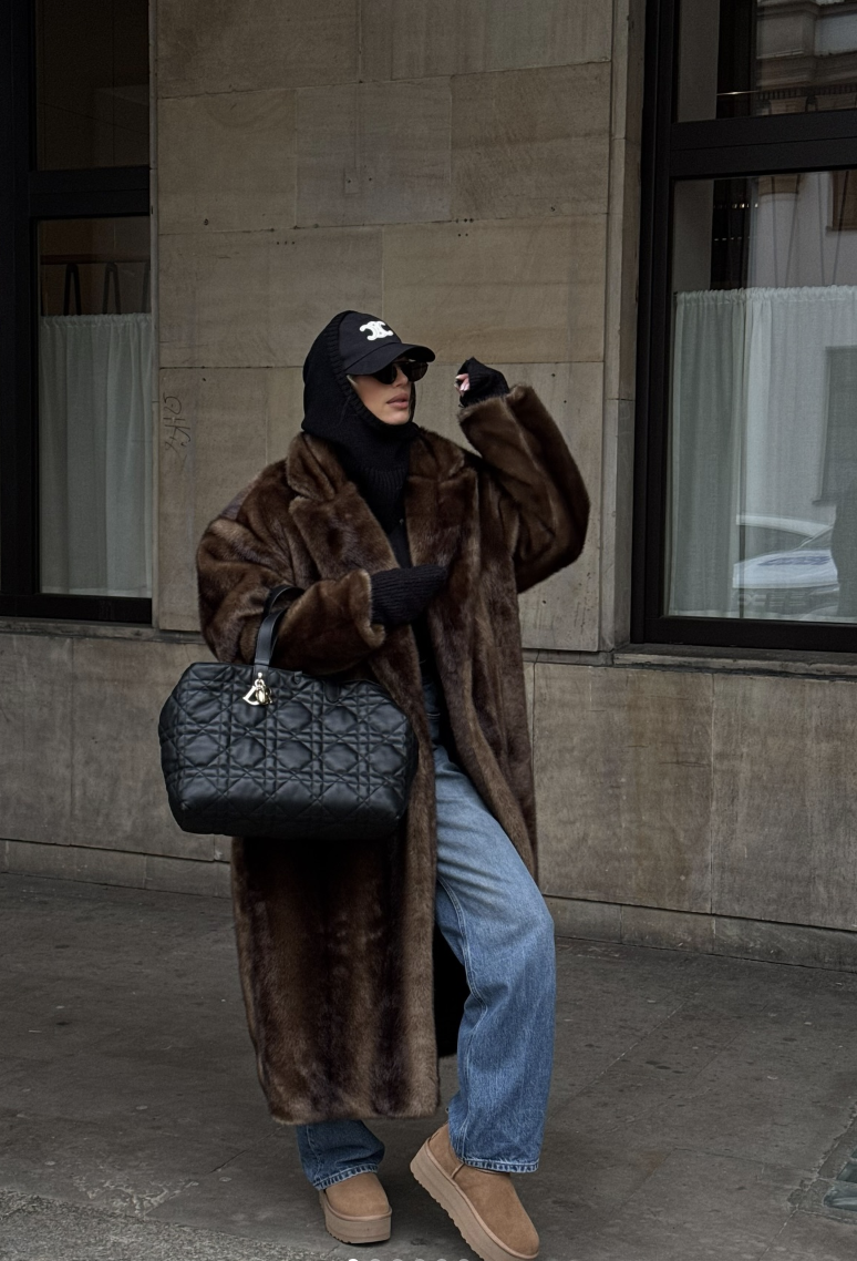 Long brown textured coat over a black top and blue jeans with tan boots and a black cap.