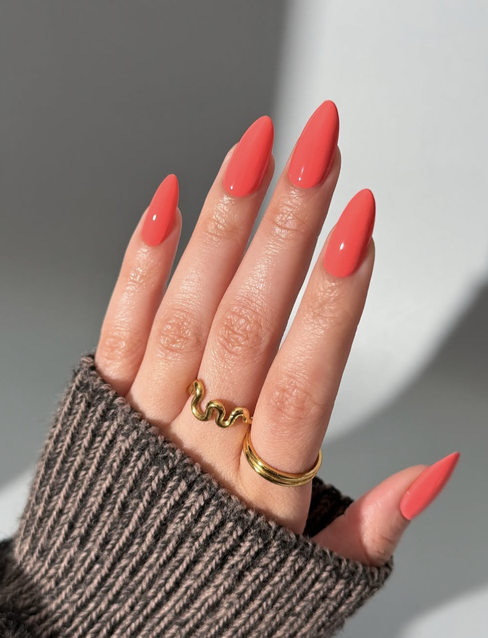 Long, almond-shaped nails showcasing a vibrant, high-gloss solid coral orange manicure accented by minimalist gold ring details.