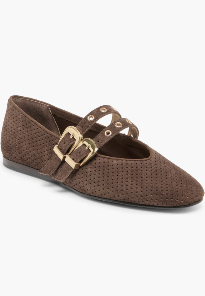 A brown suede Mary Jane flat with small grommet perforations, featuring two straps with oversized gold buckles on a transparent background.