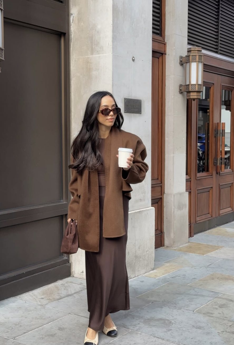 A woman wearing a chocolate brown maxi dress and matching scarf, layered under a structured camel coat with cap-toe slingback heels.