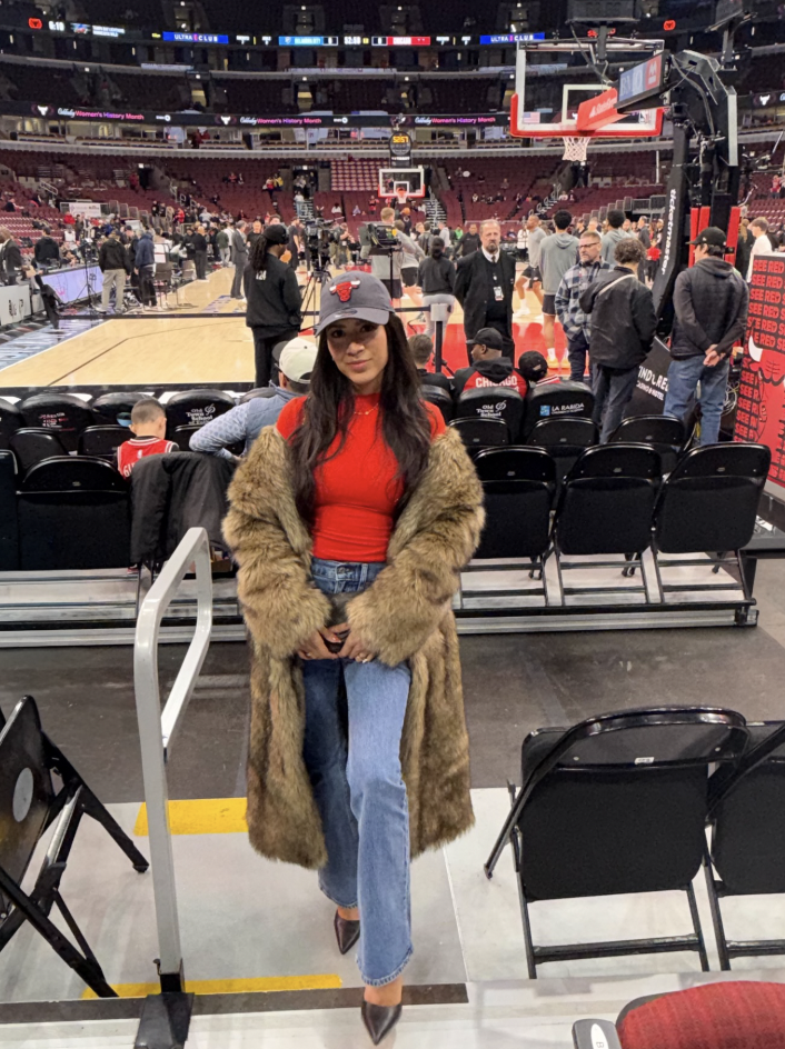 Woman courtside wearing a red fitted top, long faux-fur coat, relaxed jeans and a baseball cap.