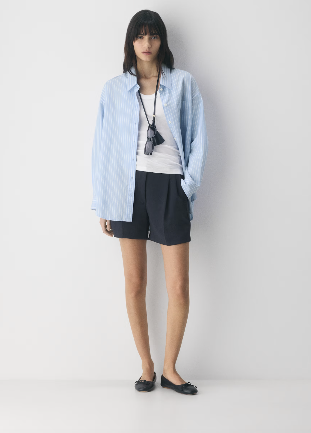 Professional traveler's pick: the Aritzia Effortless Short Mid-Thigh in black Crepette fabric, styled with black ballet flats and a light blue button-down for a wrinkle-resistant "Old Money" travel look.
