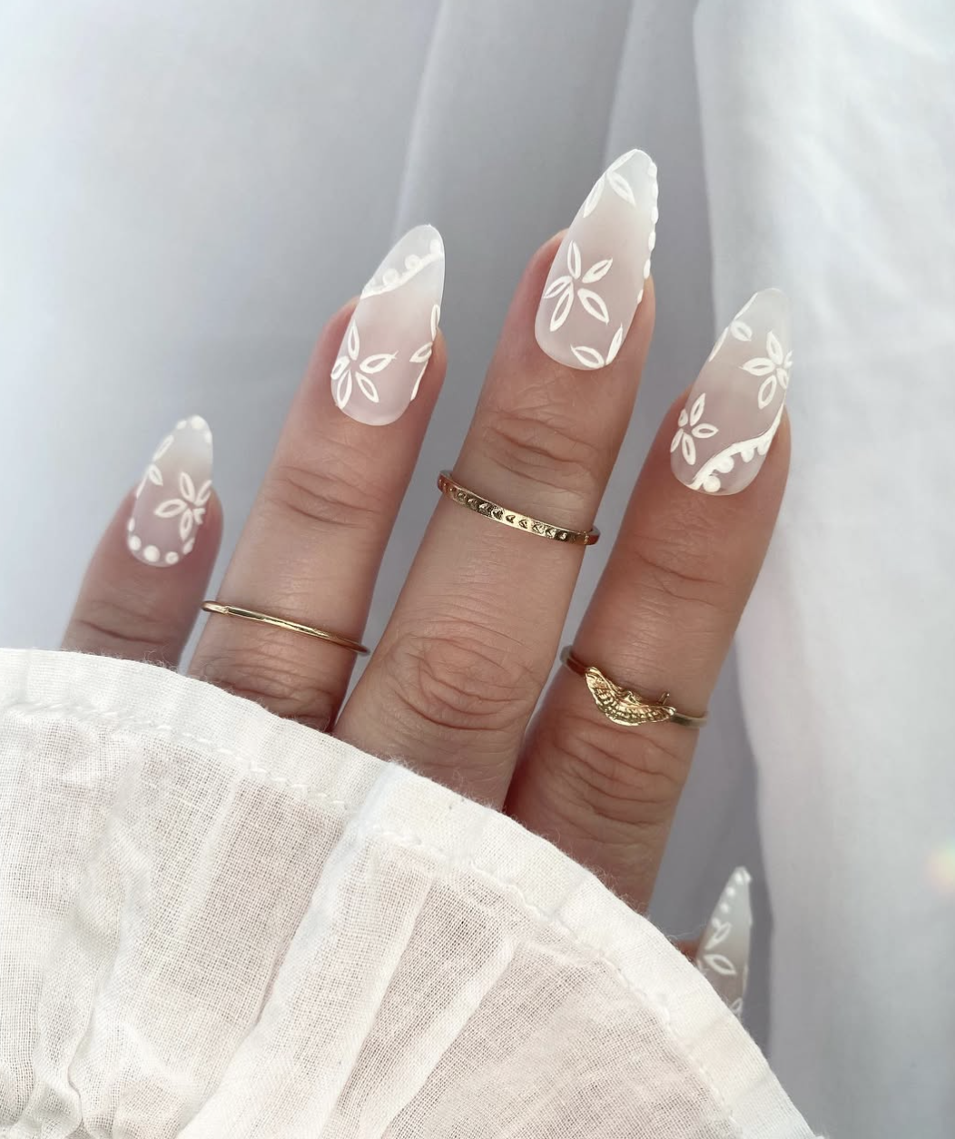 A close-up of long, almond-shaped matte milky-white nails featuring delicate hand-painted white floral outlines, paired with fine gold rings and a white textured sleeve.