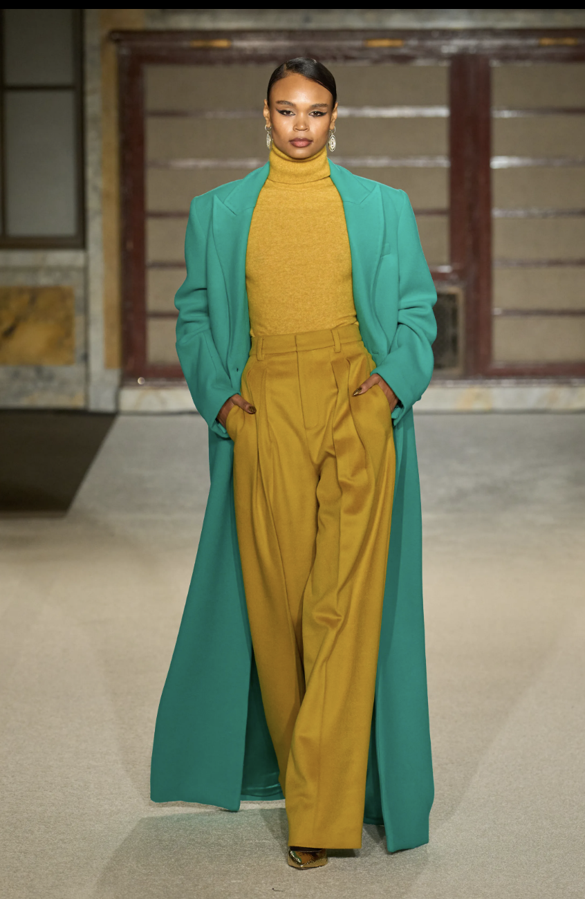 A model on the Sergio Hudson Fall/Winter 2026 runway wearing a long teal coat over mustard yellow trousers, demonstrating the aggressive color pairing trend.