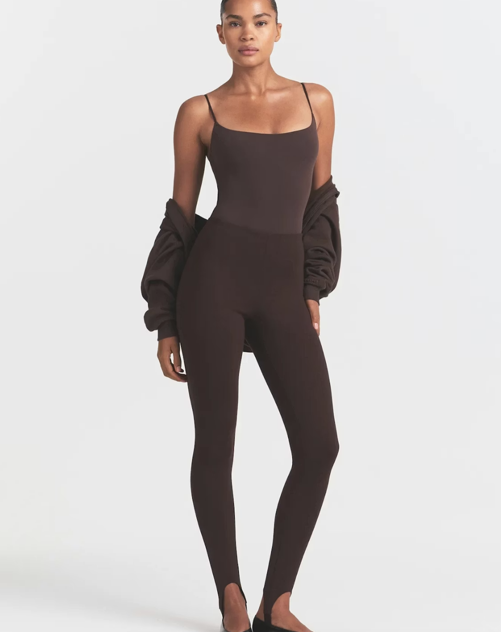 Model wearing black Skims high-waisted stirrup leggings with a matching black spaghetti strap bodysuit and a dark jacket draped over her shoulders.