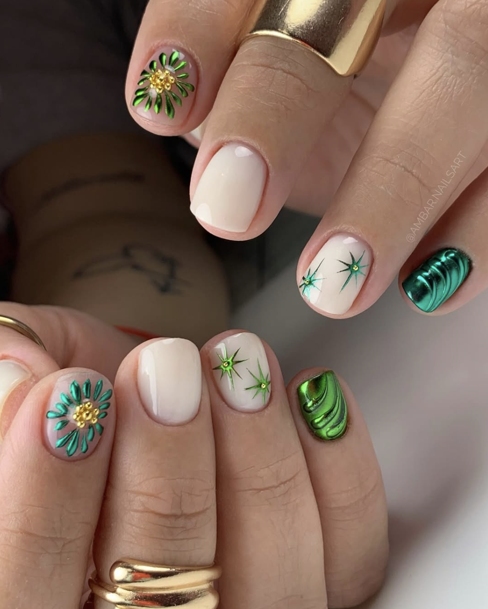 Close-up of a manicure featuring milky white base nails with 3D emerald green starburst designs, a green glitter accent nail, and structured 3D metallic green chrome waves.