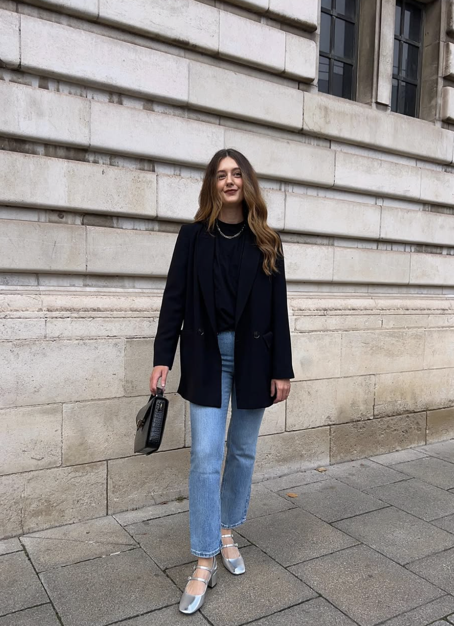 A woman standing on a city street wearing a black oversized blazer, a black tee, and cropped blue jeans paired with black Mary Jane flats for women.