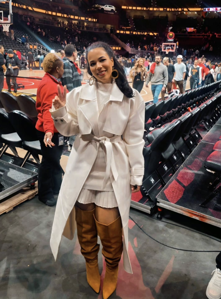 Woman courtside wearing a belted cream trench over a cream mini dress with tall cognac boots.