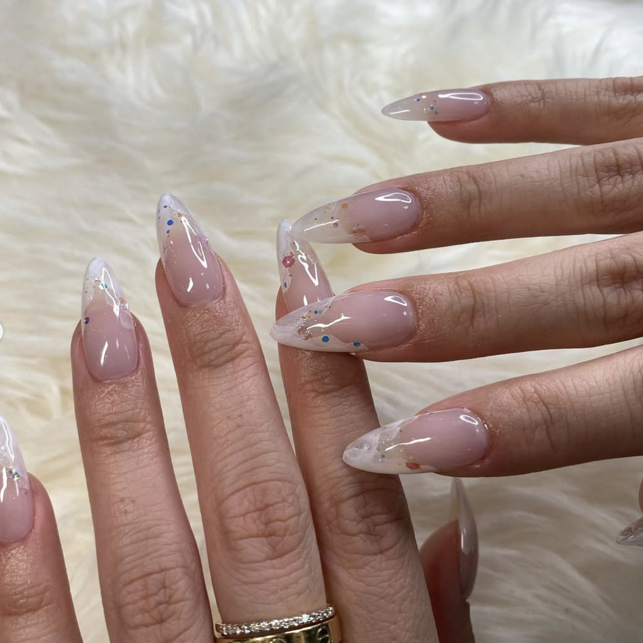 Long almond-shaped nails with a flawless, high-shine sheer nude finish and subtle multi-colored glitter accents.