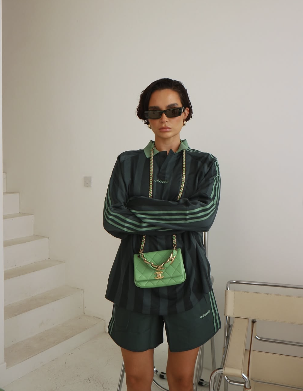 A woman wearing dark green nylon track shorts styled with a matching dark green blazer, black sunglasses, and a green shoulder bag for an elevated summer street style look.