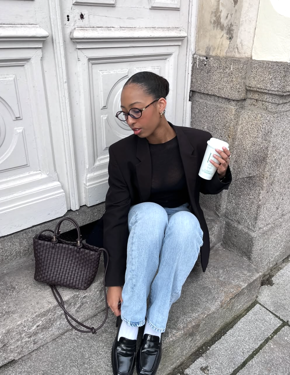 A woman sitting on a stone ledge wearing a sleek black blazer over a black satin camisole and light-wash straight-leg jeans, paired with black loafers and a woven leather handbag.