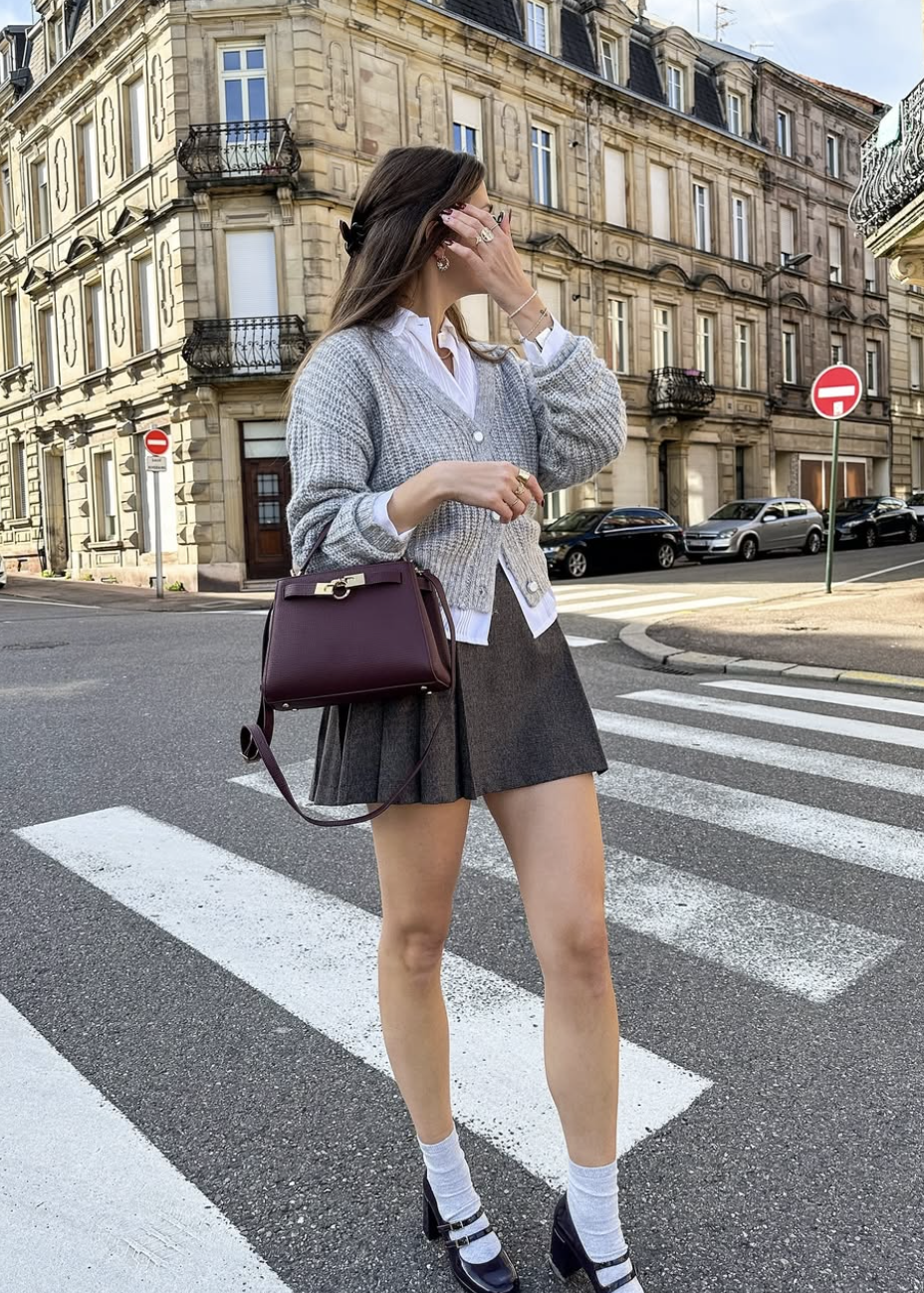 A woman walking across a city street wearing a grey cardigan over a white button-down, a dark pleated mini skirt, and black Mary Jane flats for women with white crew socks.