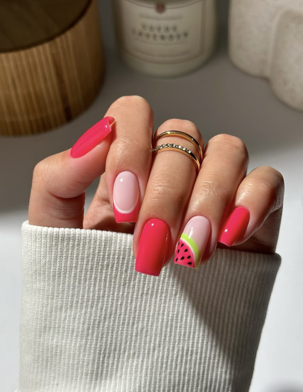 A close-up of square-shaped nails with a vibrant hot pink French manicure, featuring a playful accent nail painted with a detailed watermelon slice pattern complete with tiny black seeds, paired with a white knit sleeve.