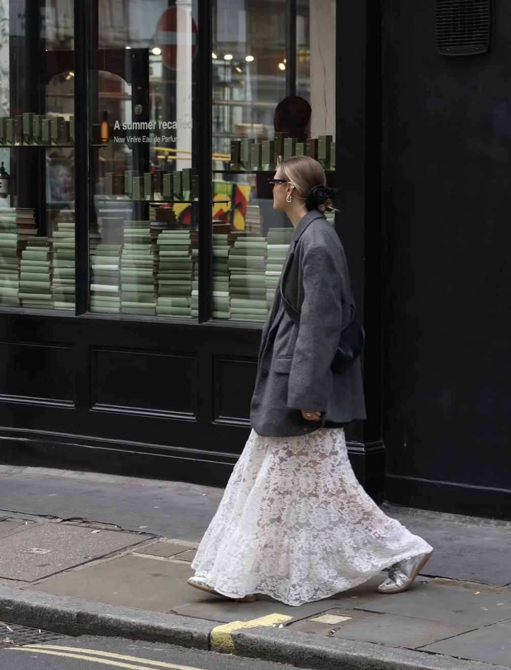 A woman walking down a city street wearing a sheer white floral lace maxi skirt paired with an oversized grey blazer and dark sunglasses.