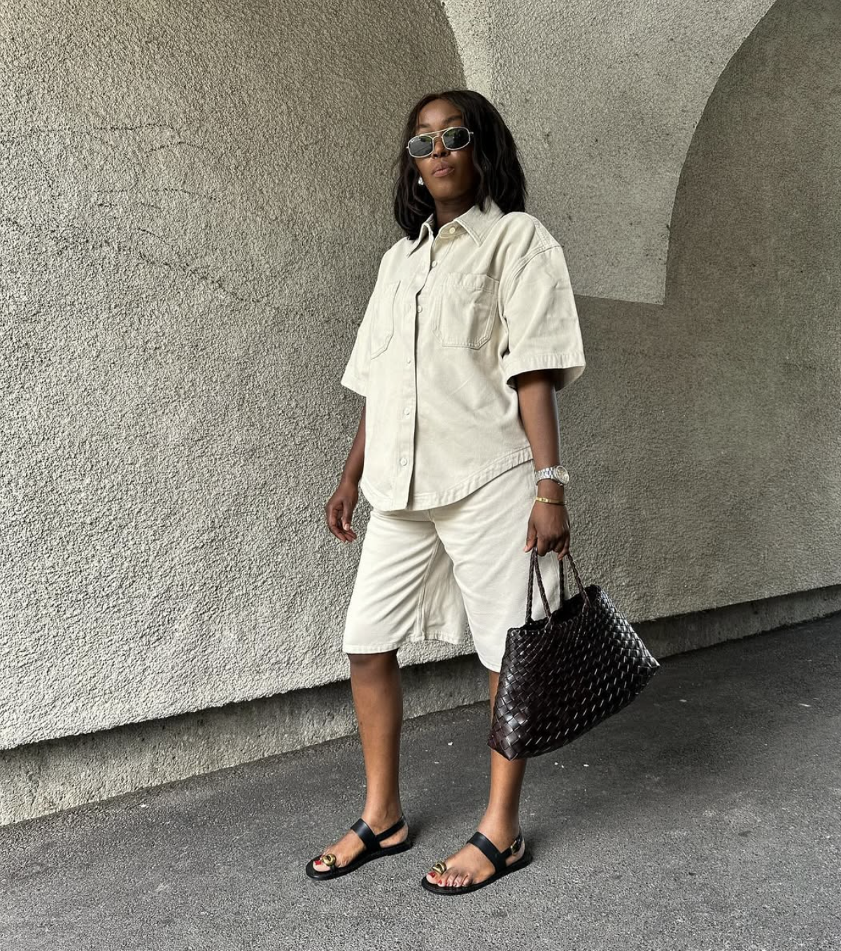 A woman wearing a matching cream-colored short-sleeved linen button-down shirt and Bermuda shorts, paired with black slide sandals and a woven black leather tote bag.