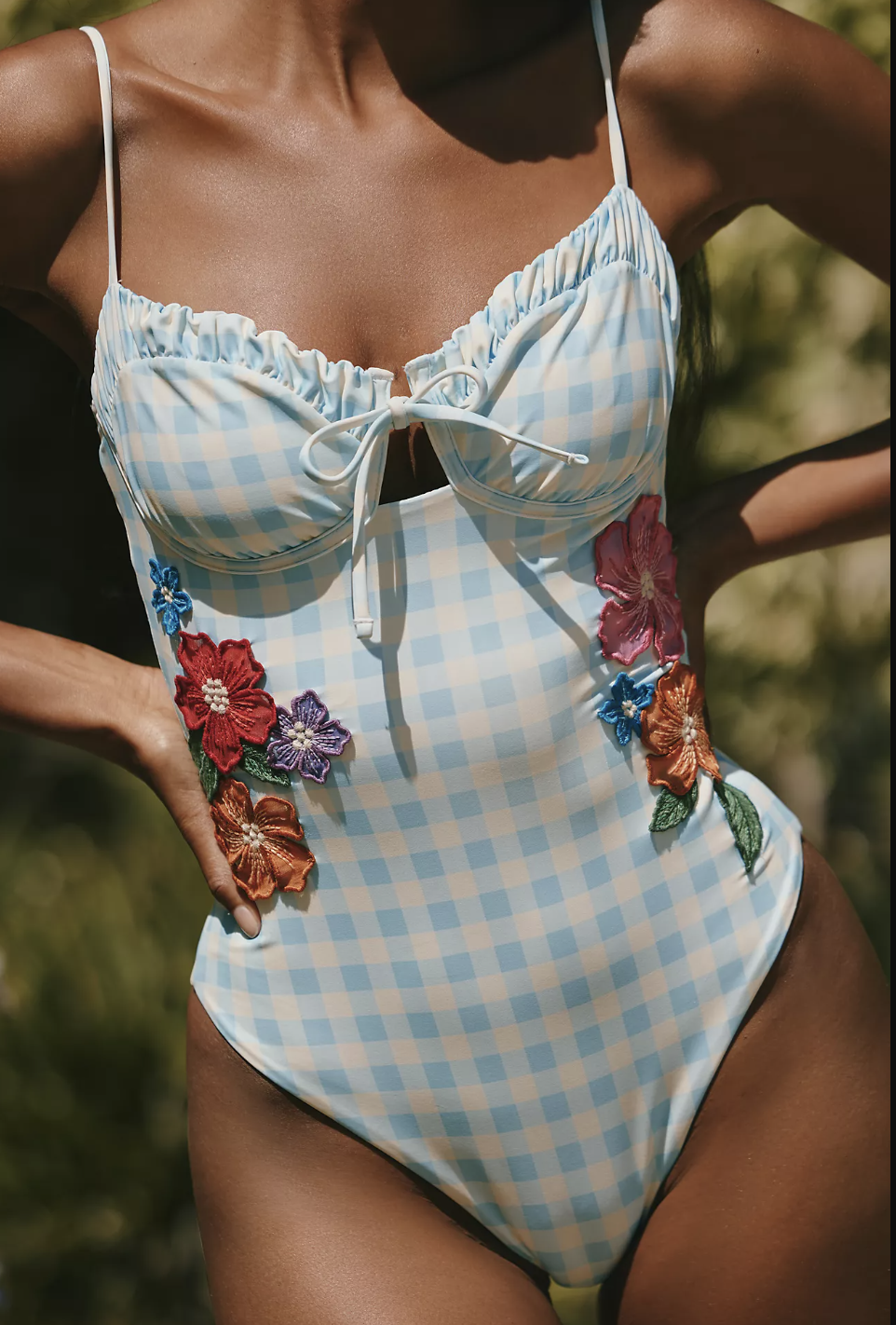 A close-up of high-waist bikini bottoms featuring a classic light blue and white gingham check pattern with a decorative orange floral accent at the hip.