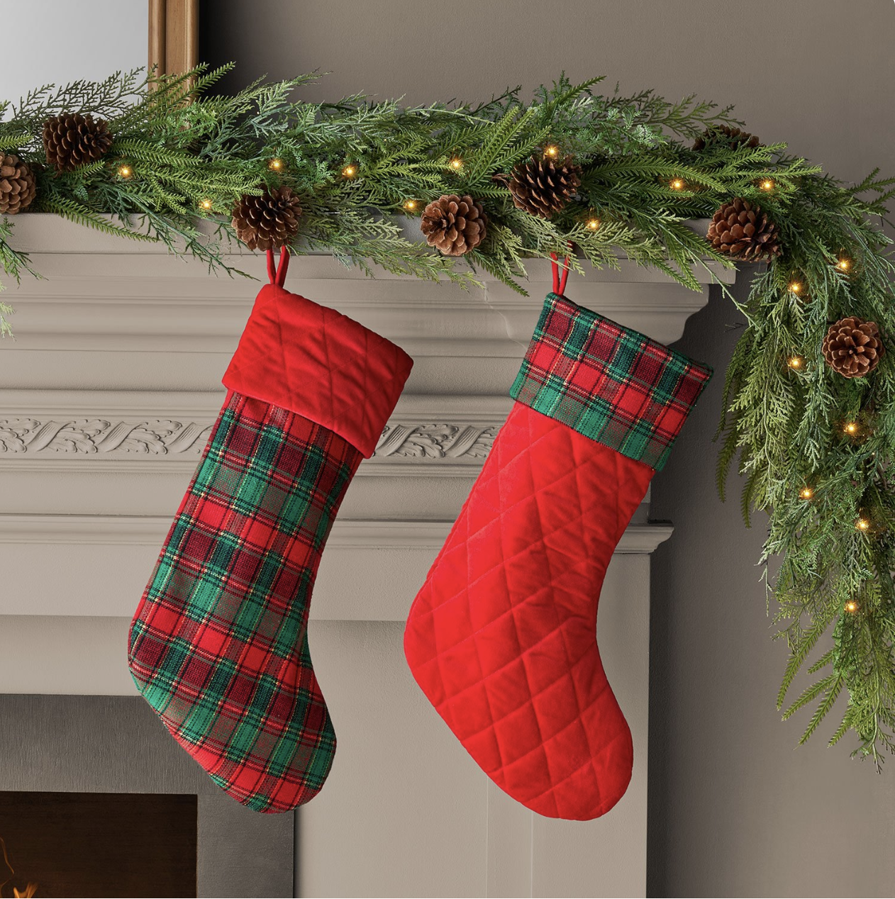 Red quilted velvet and red-and-green plaid Christmas stockings hanging under a lit cedar garland with pinecones.