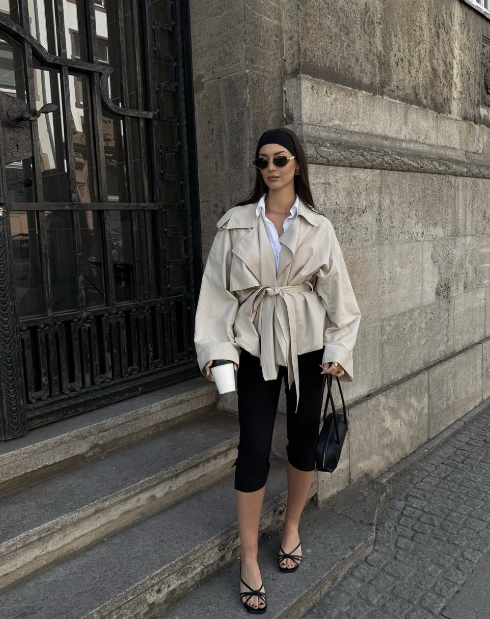 A woman wearing black capri pants styled with a beige belted utility jacket, a white button-down shirt, and black strappy heels.