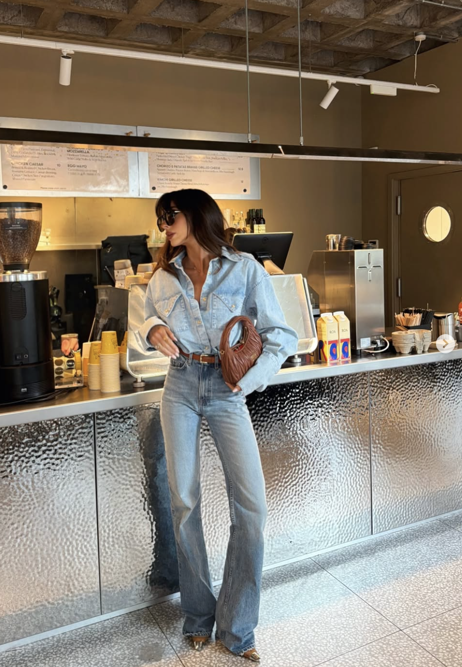 A woman standing in a cafe wearing a light-wash denim button-up shirt tuck into matching light-wash jeans, styled with a brown woven leather shoulder bag and sunglasses.