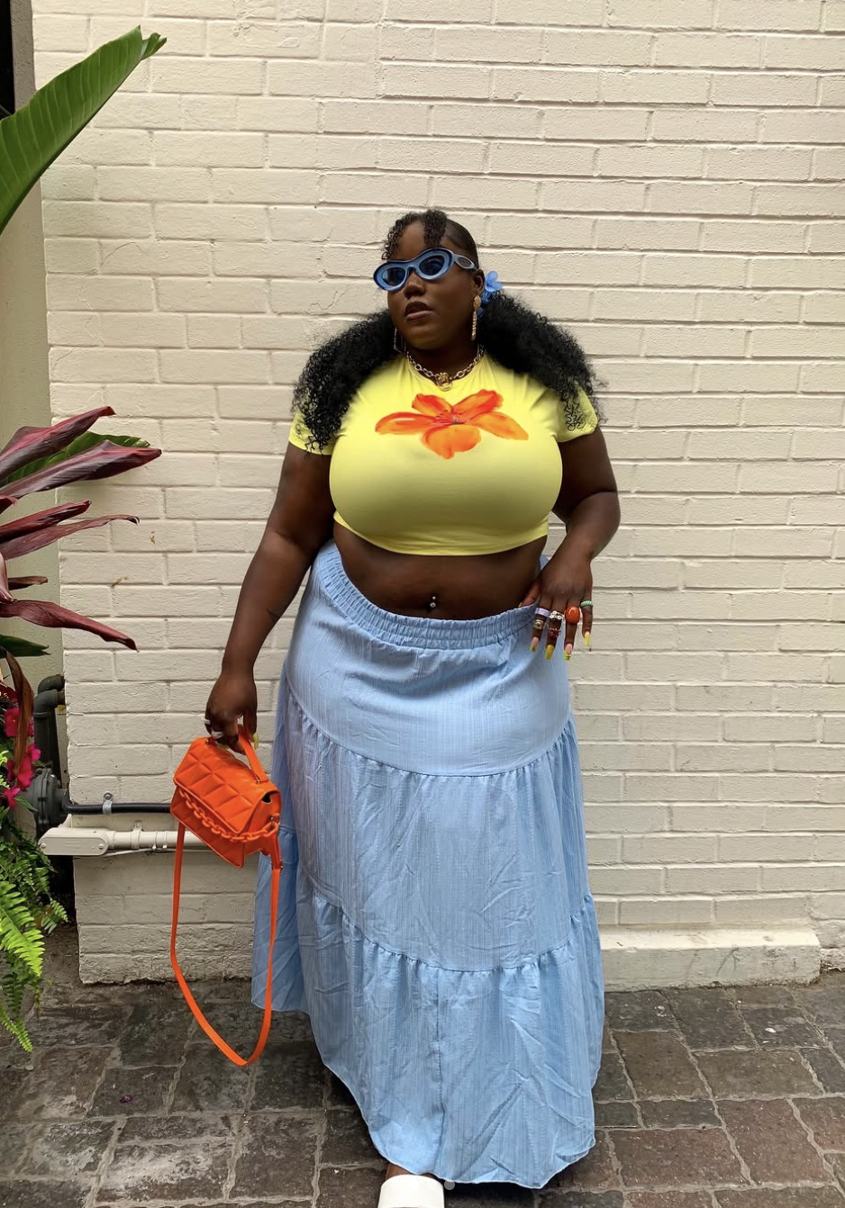 A woman wearing a neon yellow cropped t-shirt with a large vibrant orange flower graphic on the front, paired with a light blue tiered maxi skirt, and holding a bright orange quilted top-handle handbag.