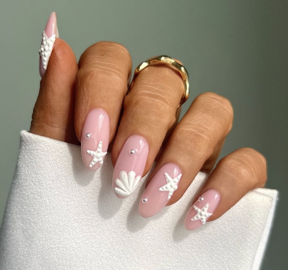 A close-up of long, almond-shaped nails featuring a sheer pink base decorated with 3D matte white mermaid-themed artwork, including detailed textures of a starfish and a scalloped seashell, accented with small pearl embellishments.