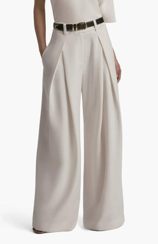A woman wearing the Favorite Daughter "The Favorite Pant" in a beige pleated wide-leg silhouette, styled with a black belt—a foundational piece for an office capsule wardrobe.