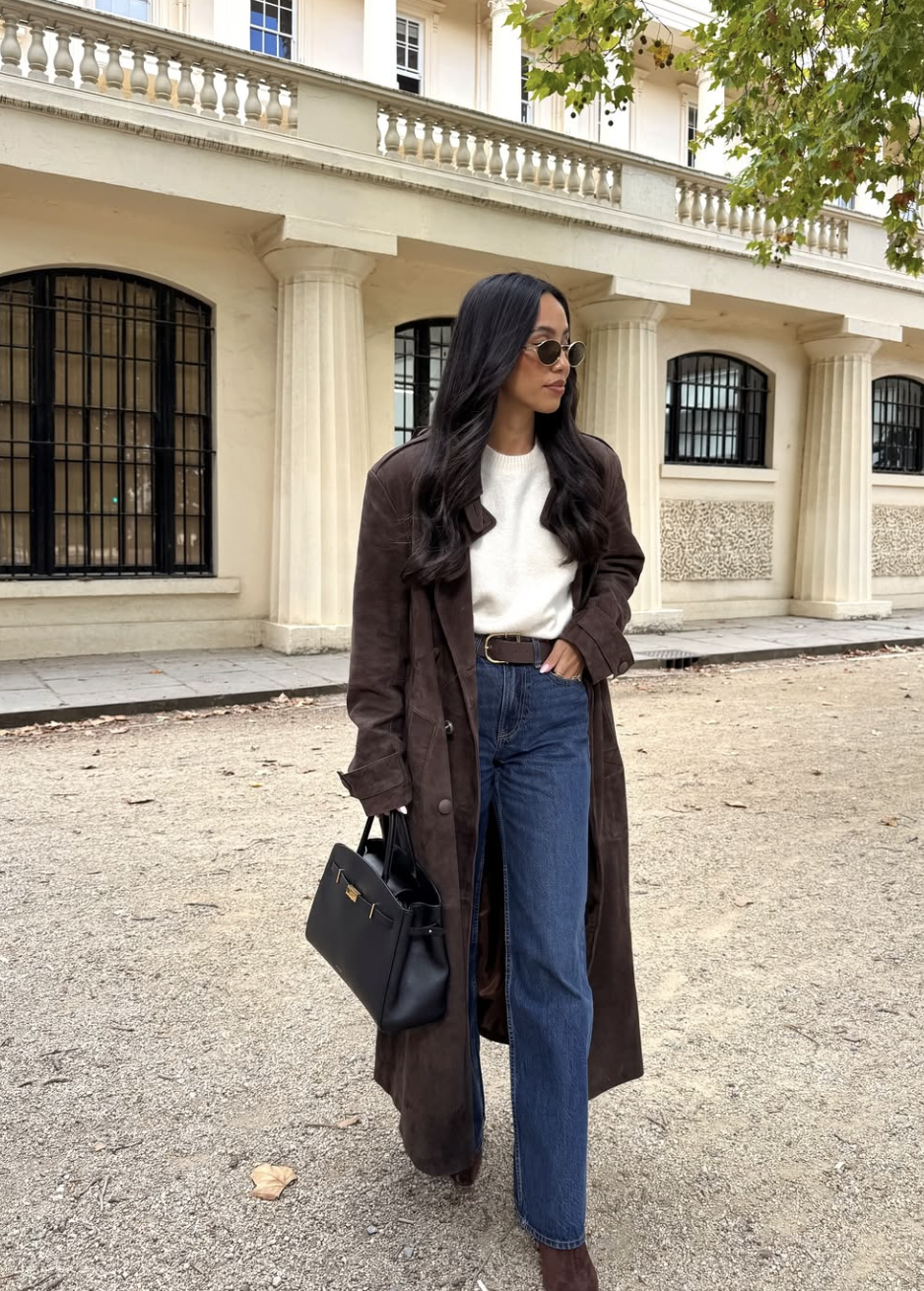 A woman wearing a long chocolate brown trench coat over a white t-shirt and dark-wash wide-leg jeans, styled with a black leather belt and sunglasses.