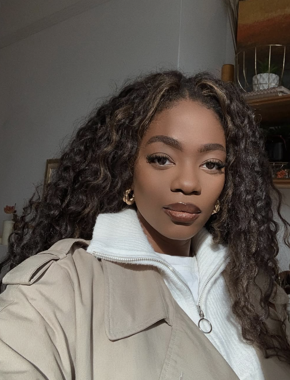 A woman with voluminous, curly crochet braids featuring a side part, wearing a tan trench coat over a white zip-up sweater and gold hoop earrings.