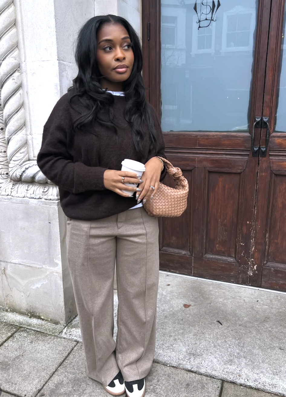 A woman wearing an oversized dark brown sweater and tan trousers with front seams, carrying a tan woven shoulder bag.