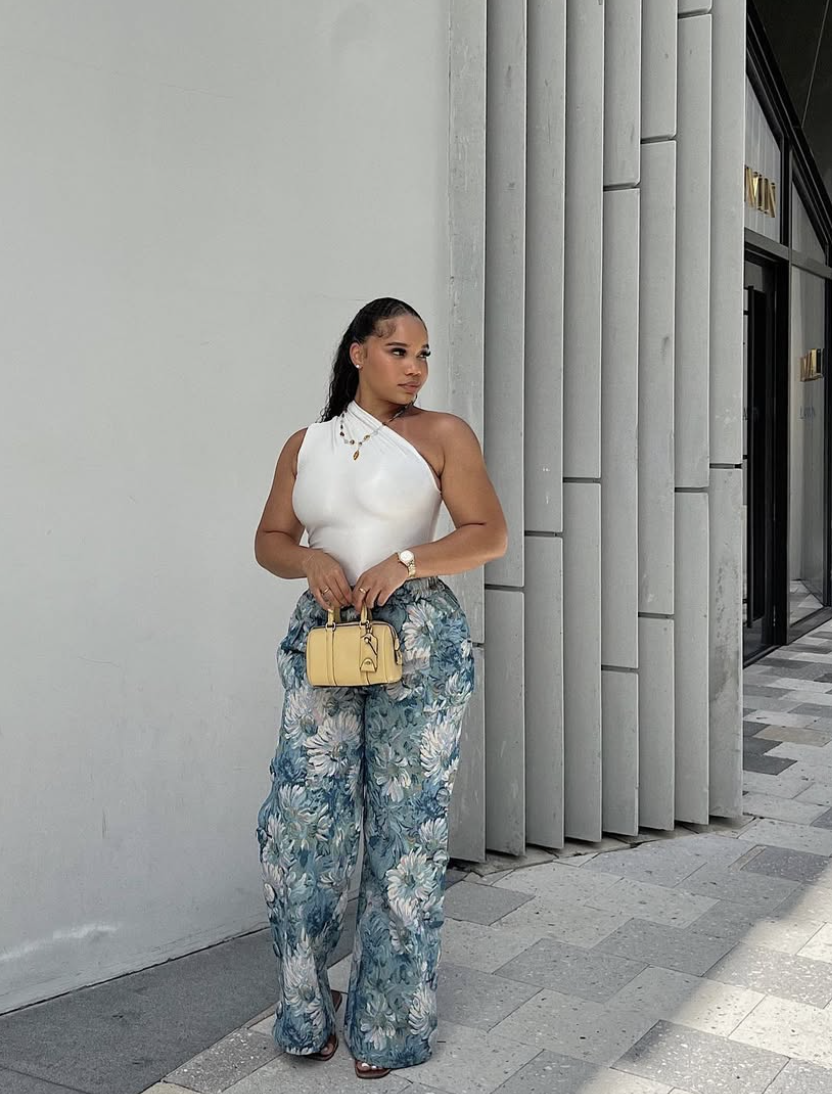 A woman wearing a white sleeveless ribbed top and blue and white floral patterned wide-leg linen pants, carrying a yellow handbag and standing against a modern grey slatted wall.