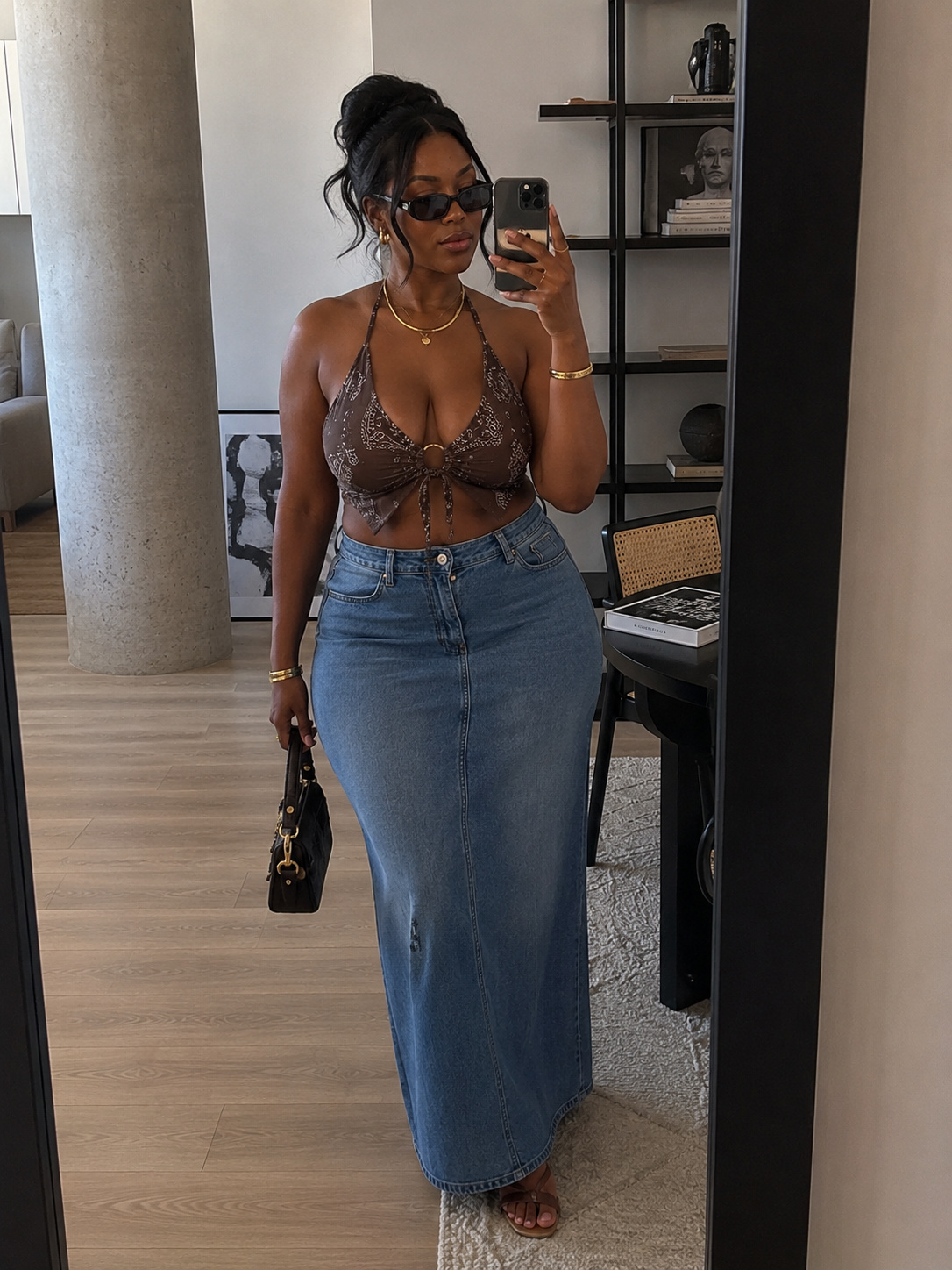 A woman with her hair in an updo wearing sunglasses, a brown knit bralette top, and a long denim maxi skirt.