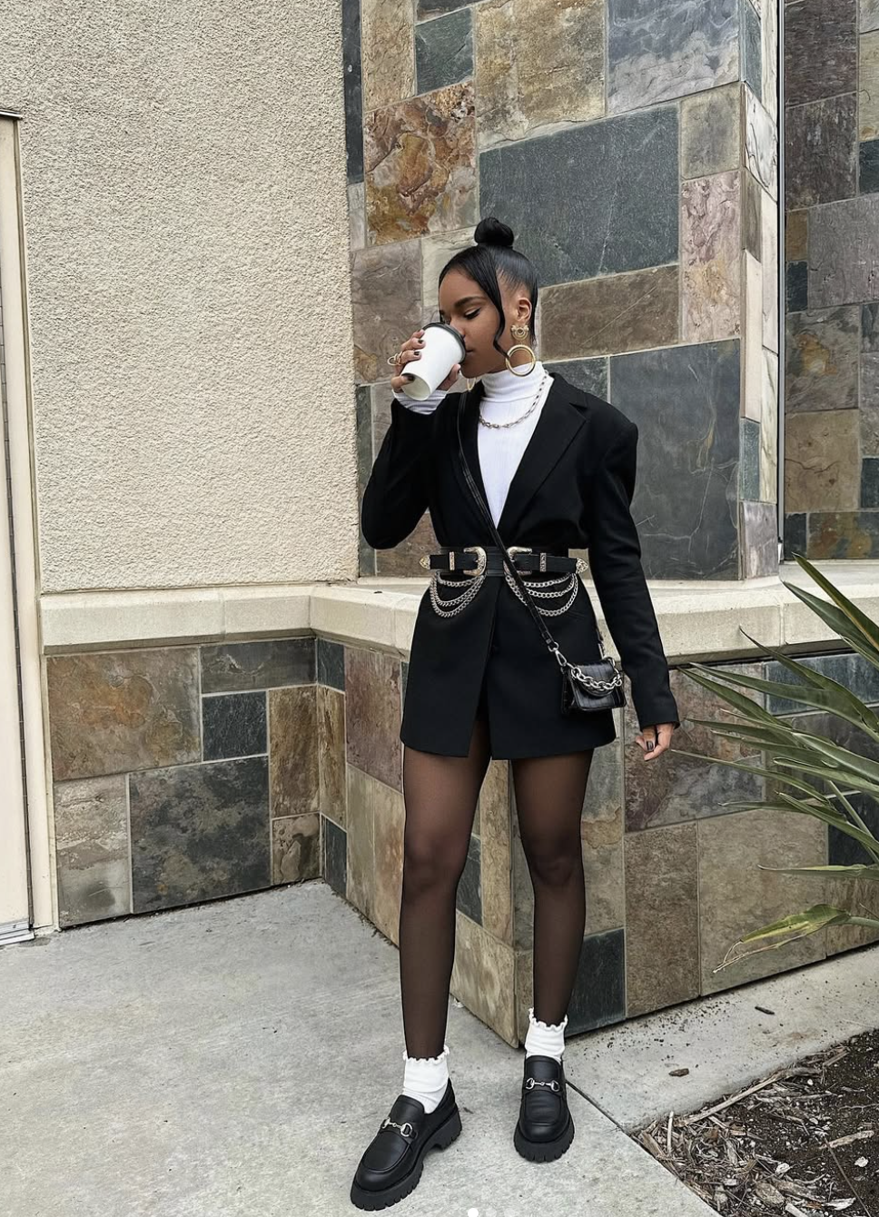 A woman wearing an oversized black blazer cinched with a double-chain belt, paired with black tights and chunky loafers for a structured silhouette.