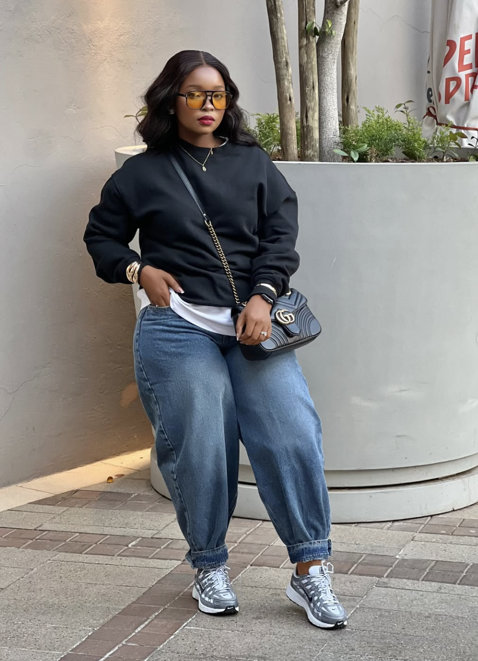 A person wearing relaxed blue barrel-leg jeans, a black crewneck sweatshirt with a white undershirt showing, metallic silver sneakers, and a black Gucci quilted crossbody bag.