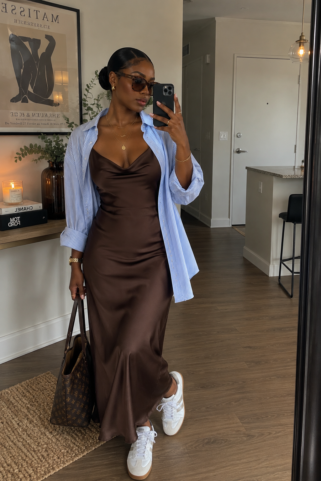 A woman styling a rich brown satin slip dress layered with an open, oversized blue-and-white striped button-down shirt and clean white sneakers.