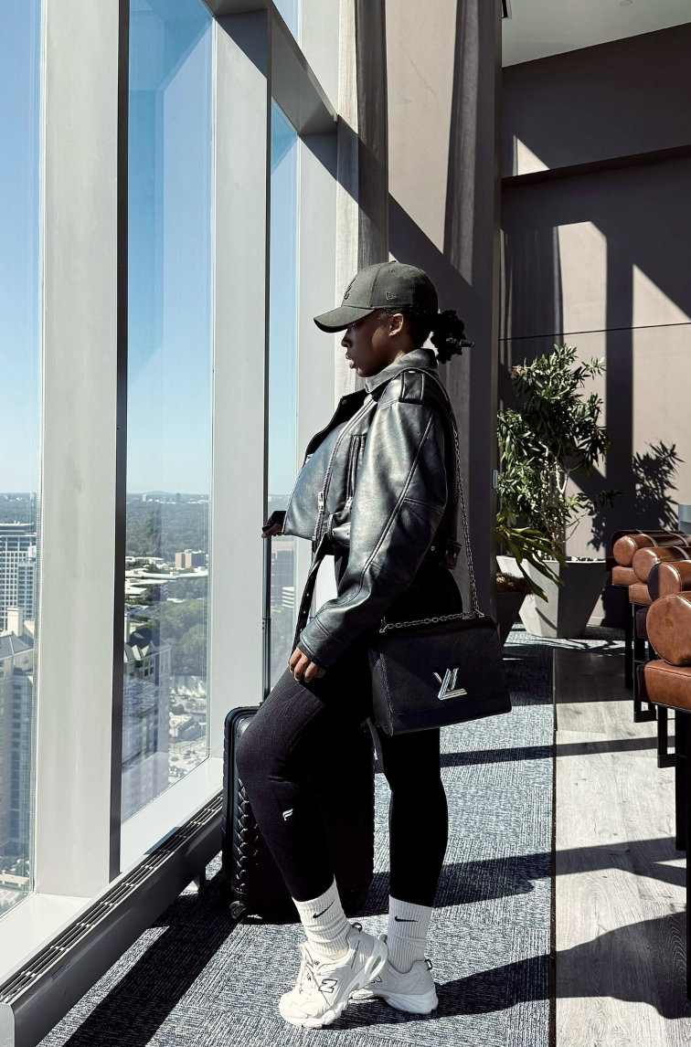 Black leather-look jacket and black leggings with white crew socks, white sneakers, an olive cap and a compact crossbody.