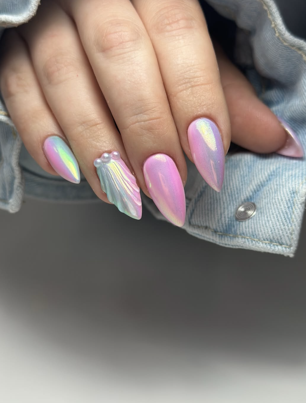 Long pointed almond nails with a mesmerizing pink and green pearlescent mermaid chrome finish, featuring a 3D textured seashell accent nail adorned with tiny pearls, highlighting cute June nail ideas.