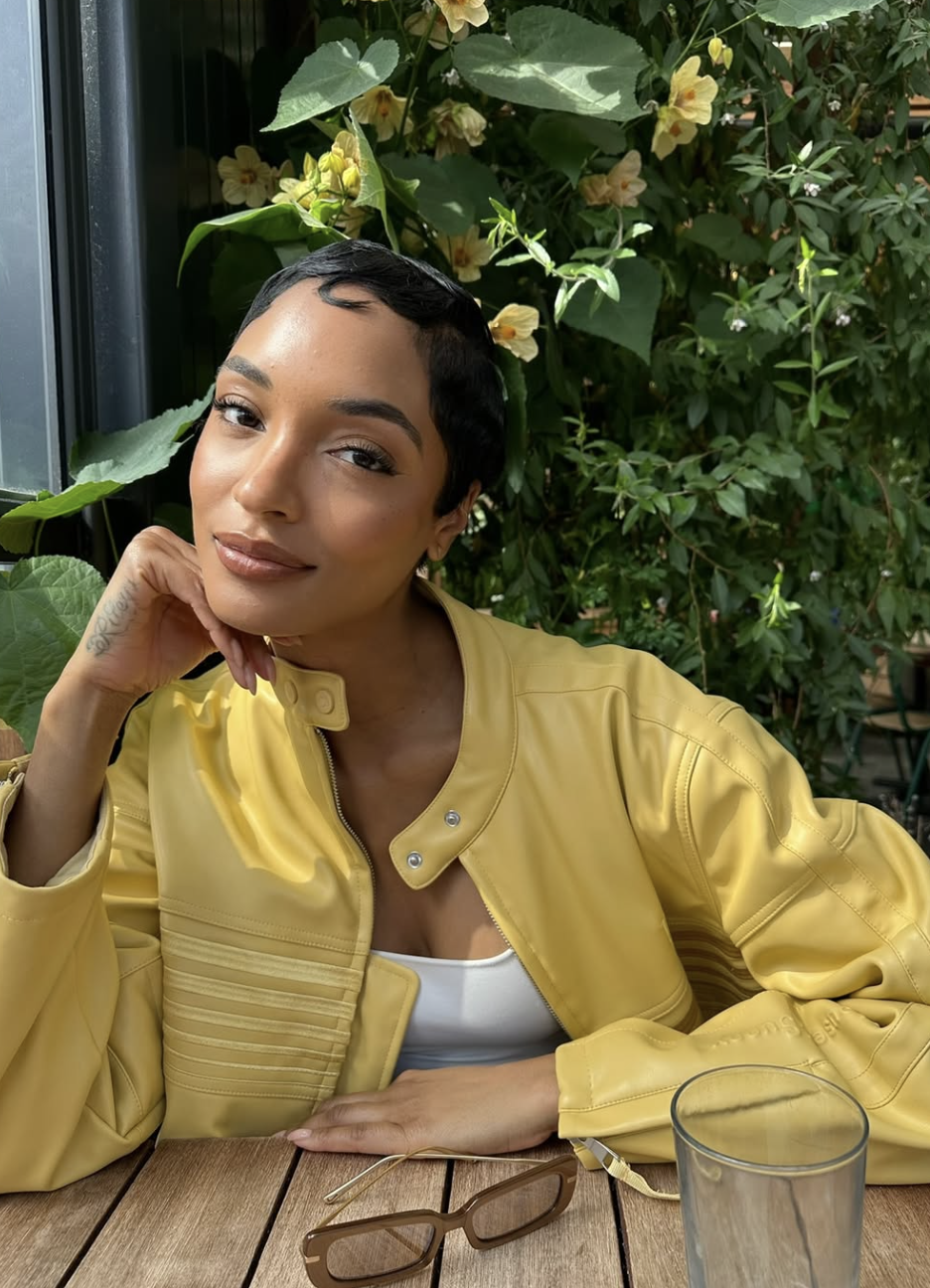 A woman with an oval face shape featuring a sleek, side-parted pixie cut with styled edges, wearing a yellow leather jacket.