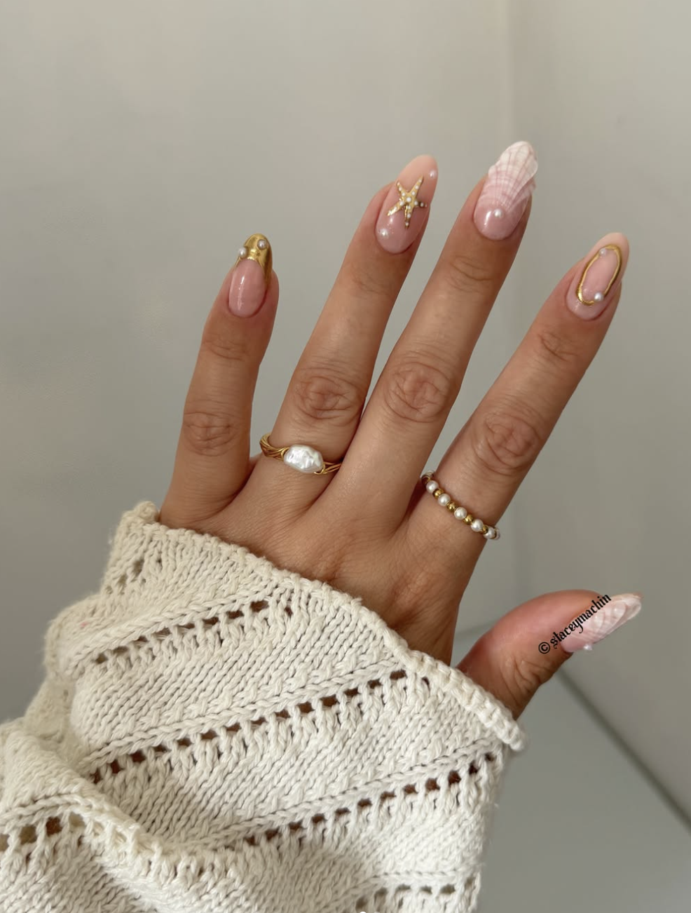 A close-up of almond-shaped nails featuring a minimal nude base with abstract 3D gold wire accents outlining the tips and cuticles, accessorized with pearl and baroque pearl rings against a white knit sleeve.