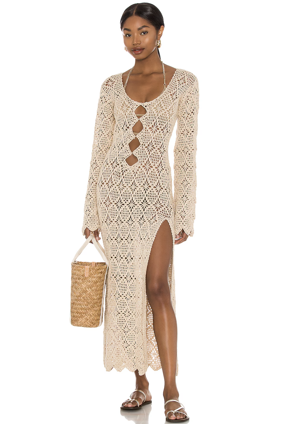 A woman wearing a beige crochet maxi dress with a front cutout, long sleeves, and a high side slit, paired with a woven straw tote bag.