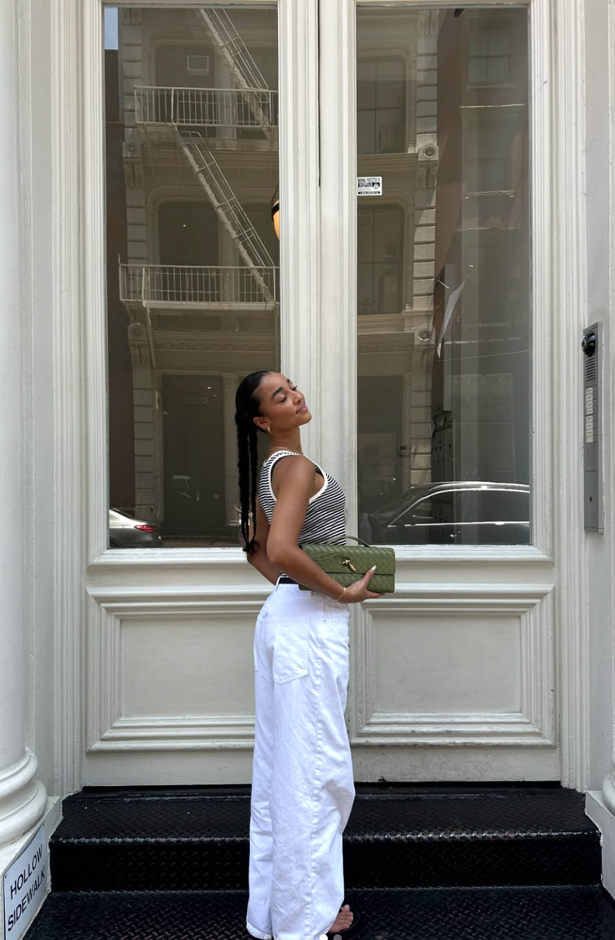 A woman standing on city stairs wearing white straight-leg jeans, a black and white striped tank top, and carrying a green textured clutch bag.