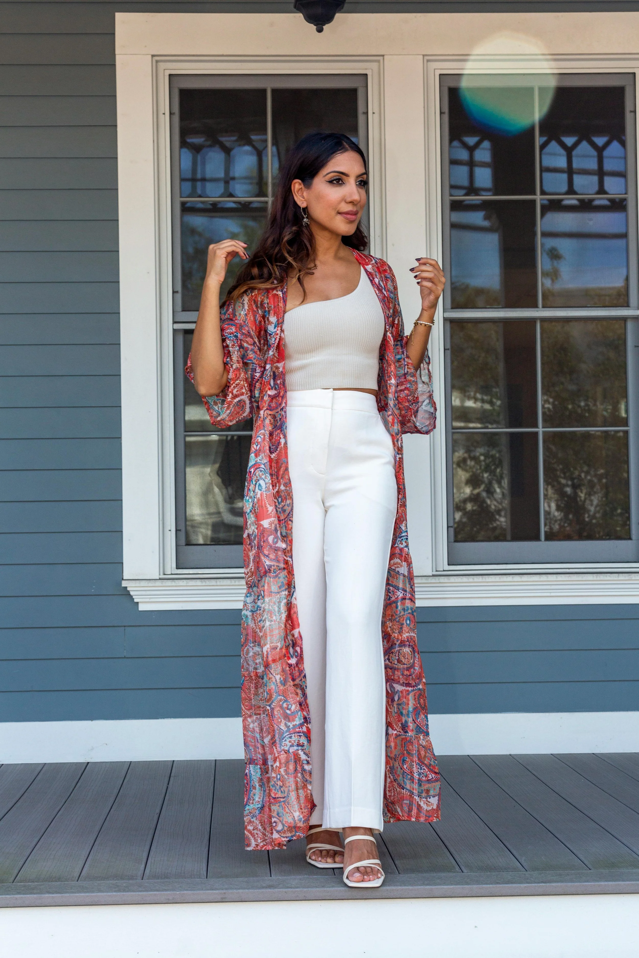 A woman standing on a deck wearing a long, colorful floral print kimono over a white top and wide-leg white trousers with tan sandals.