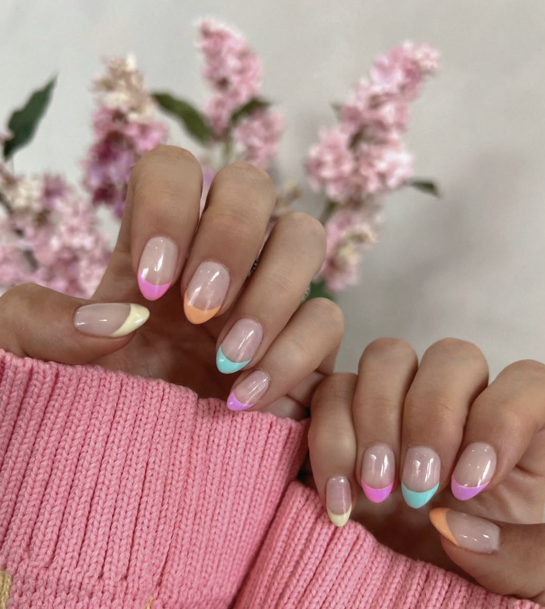 A close-up of a hand with medium almond-shaped nails featuring a sheer nude base and thick "ribbon-style" French tips in mismatched pastel colors including pink, orange, green, and purple.
